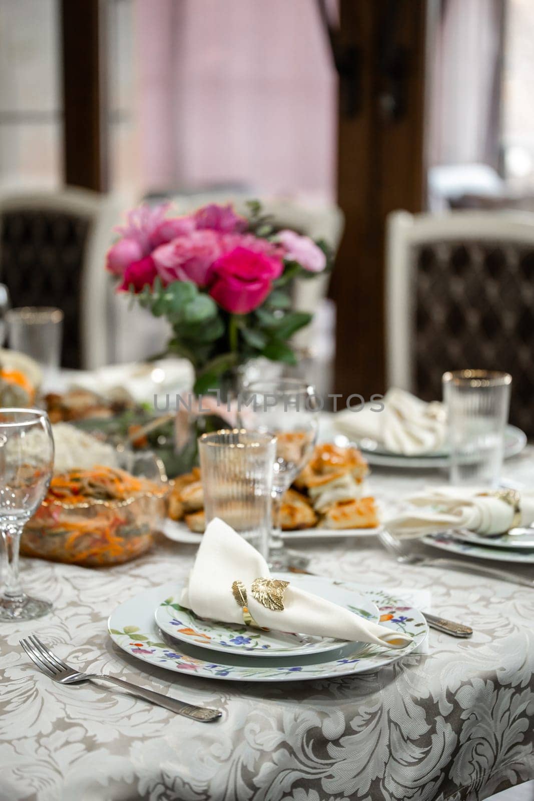 An elegant table setting with pink roses centerpiece in a crystal vase, fine china plates, silverware, and crystal wine glasses. White tablecloth, blurred background with window and light.