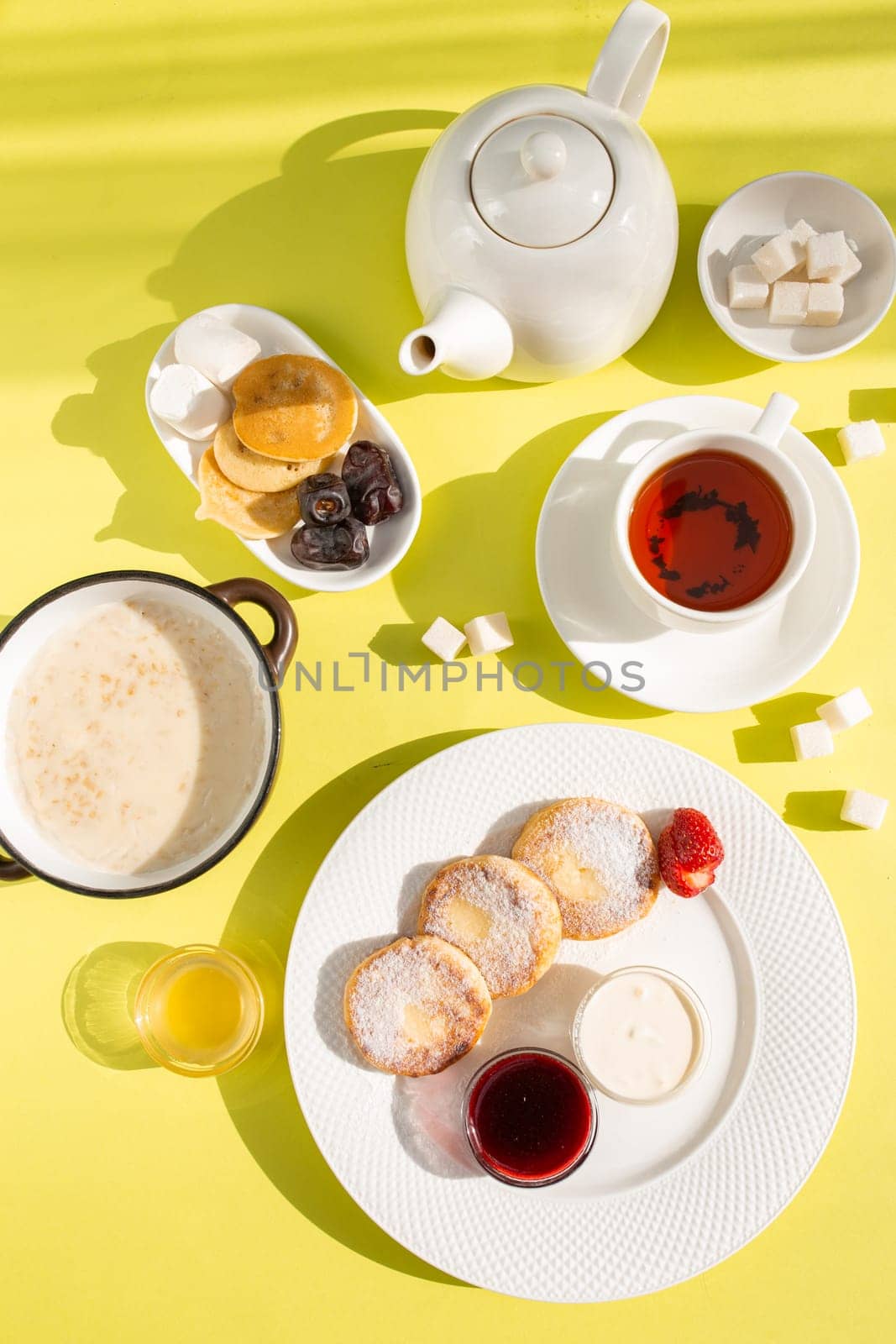 Savor a delightful breakfast with cottage cheese pancakes, creamy oatmeal, aromatic tea, golden honey, jam, and fresh fruit on a sunny yellow table, creating a vibrant start to your day.