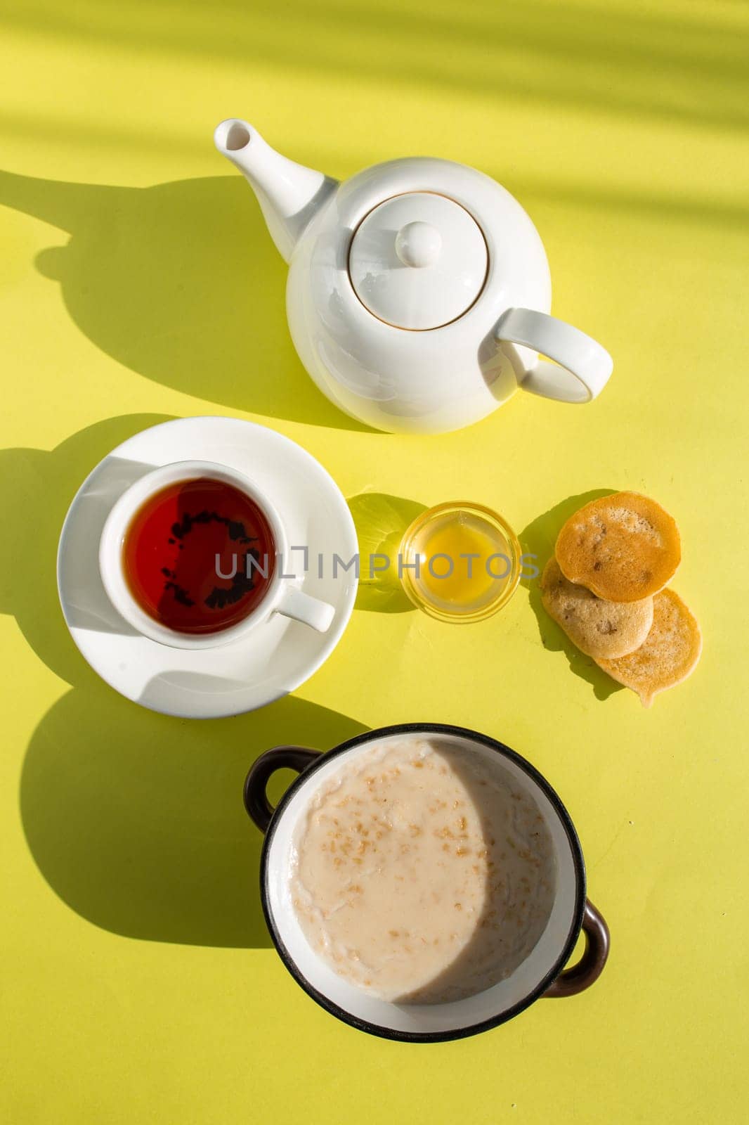 A cozy breakfast setup featuring oatmeal with milk, tea in a white cup on saucer, honey in a glass bowl, and bread slices on a yellow background. Shadows add depth and warmth to the scene.