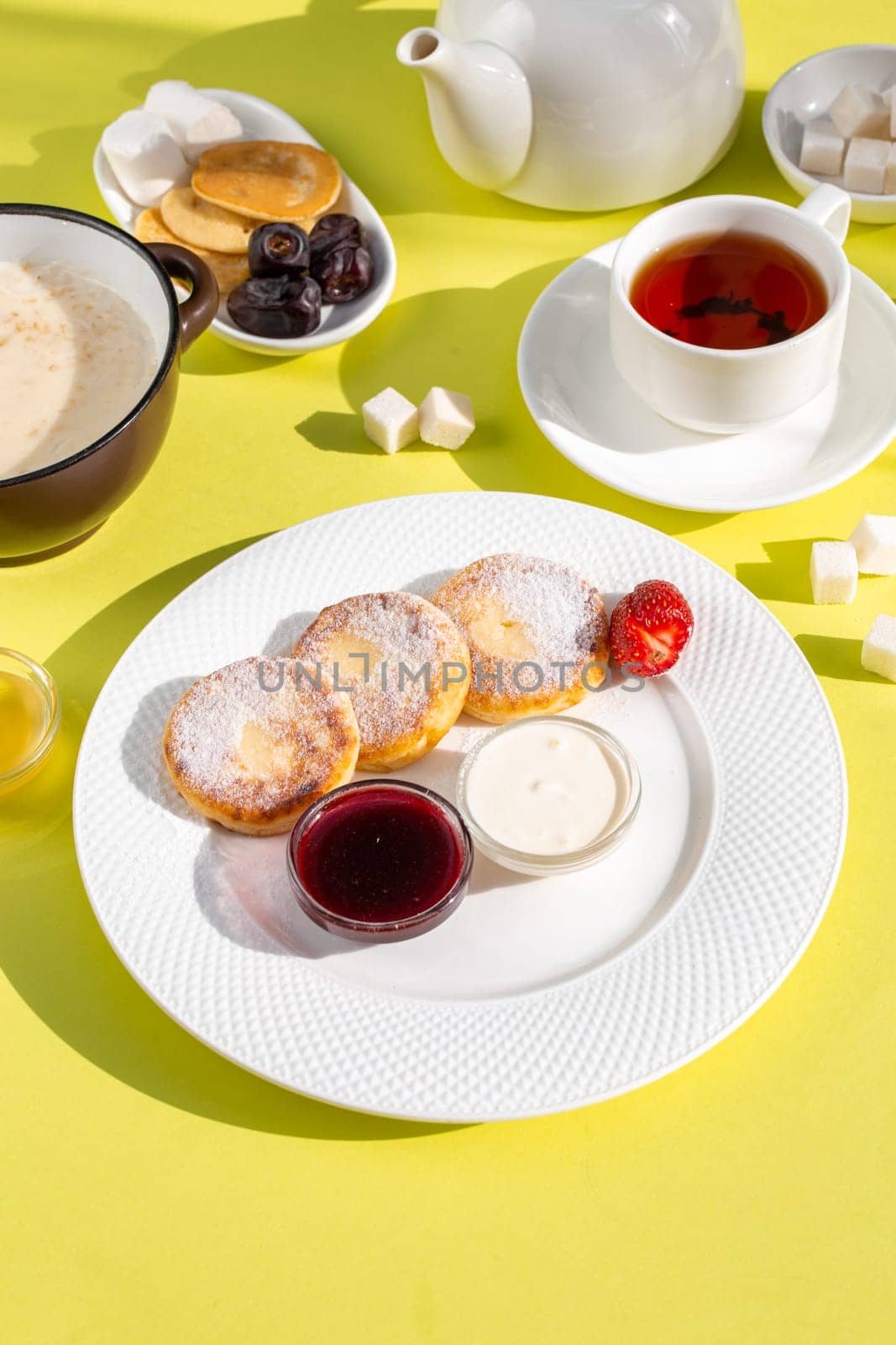 Savor a delightful breakfast with cottage cheese pancakes, creamy oatmeal, aromatic tea, golden honey, jam, and fresh fruit on a sunny yellow table, creating a vibrant start to your day.