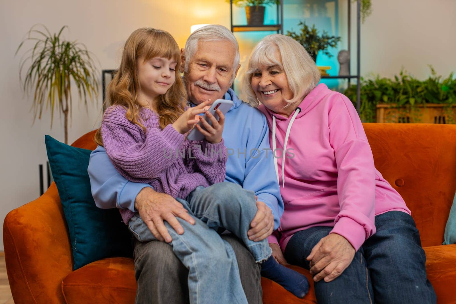 Child girl granddaughter teaching grandfather and grandmother using smartphone on sofa in home room by efuror