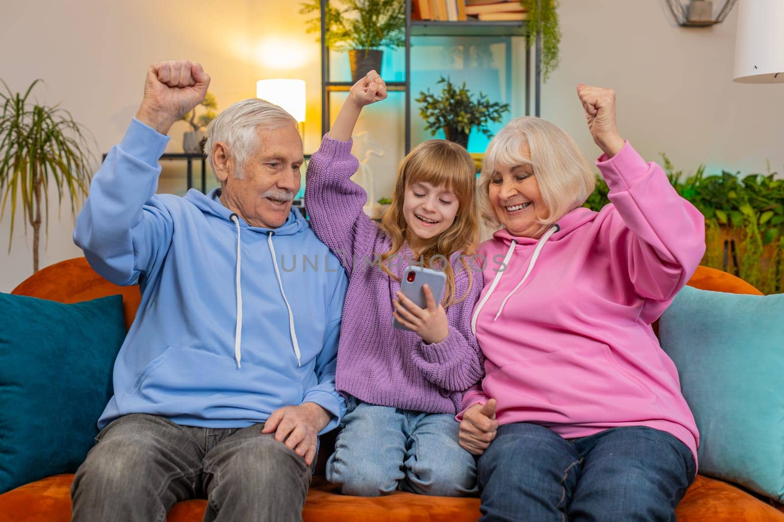 Surprise Caucasian grandfather, grandmother and granddaughter using smartphone while celebrating success at home. Happy girl with grandparents amazed by online game win sitting on sofa in living room.