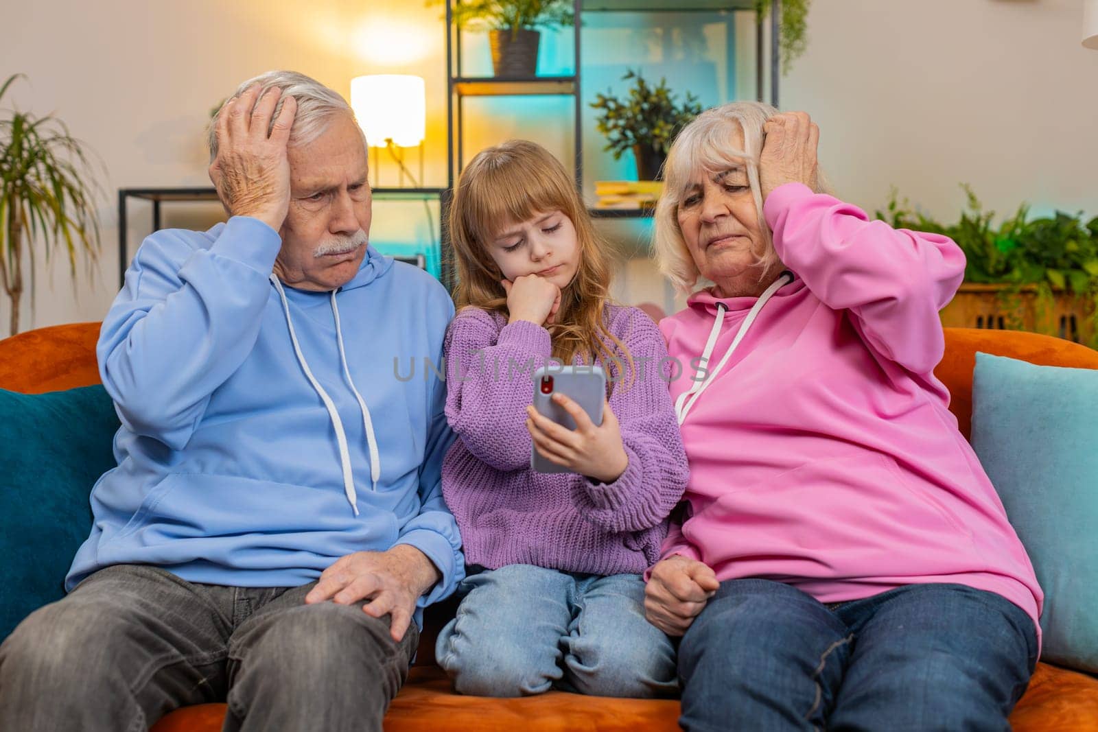 Upset grandfather, grandmother and granddaughter playing game on smartphone and losing at home. Sad Caucasian girl with grandparents received bad news, fortune loss, fail education study test on couch