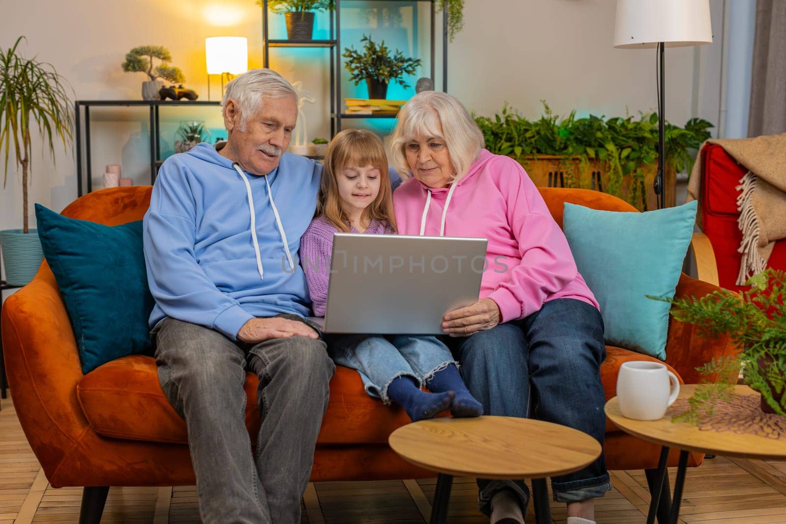 Senior Caucasian couple man and woman with child girl granddaughter spending time at home together, sitting on couch in the living room using laptop computer. Watching videos playing games on weekends