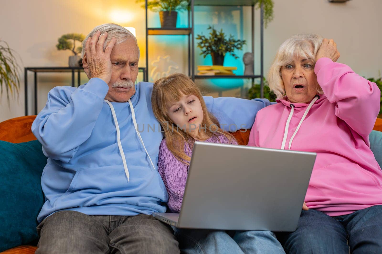 Upset senior grandfather grandmother and granddaughter playing game on laptop and losing at home. Sad Caucasian girl with grandparents received bad news fortune loss fail education study test on sofa