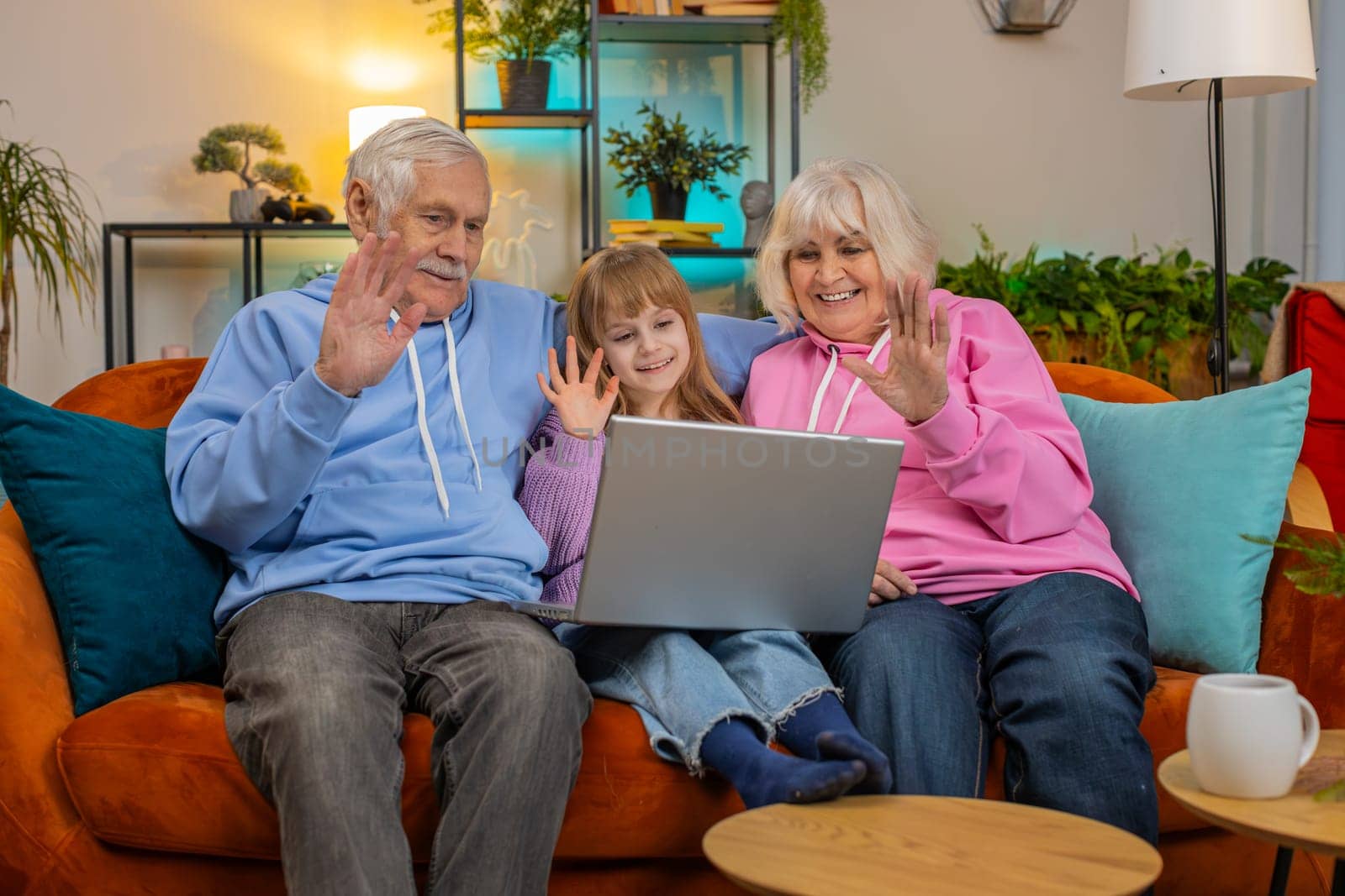 Caucasian grandfather, grandmother and granddaughter making video call online on laptop at home. Girl with grandparents talking by internet webcam chat app sitting on sofa in living room apartment.