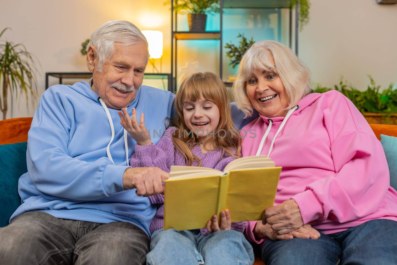 Child girl granddaughter with grandparents reading interesting book together sitting on home couch by efuror