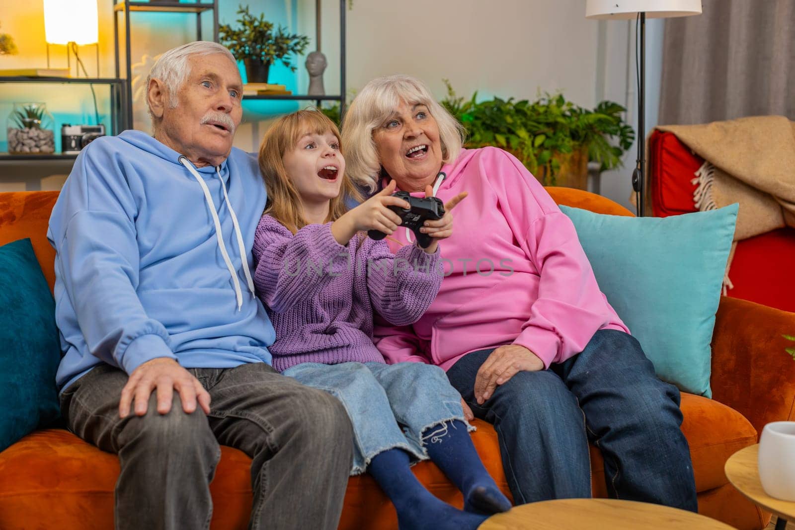 Happy Caucasian grandparents and granddaughter using joystick controller playing videogame fun on couch at home. Girl with senior man and woman enjoying success winning online game during weekends