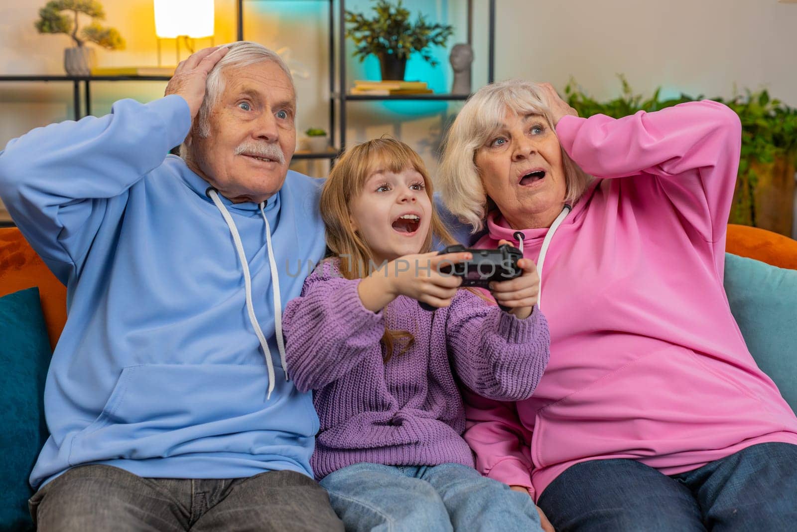 Amazed Caucasian grandparents and granddaughter using joystick controller playing videogame fun on sofa at home. Girl with senior man and woman enjoying success winning online game during weekend.