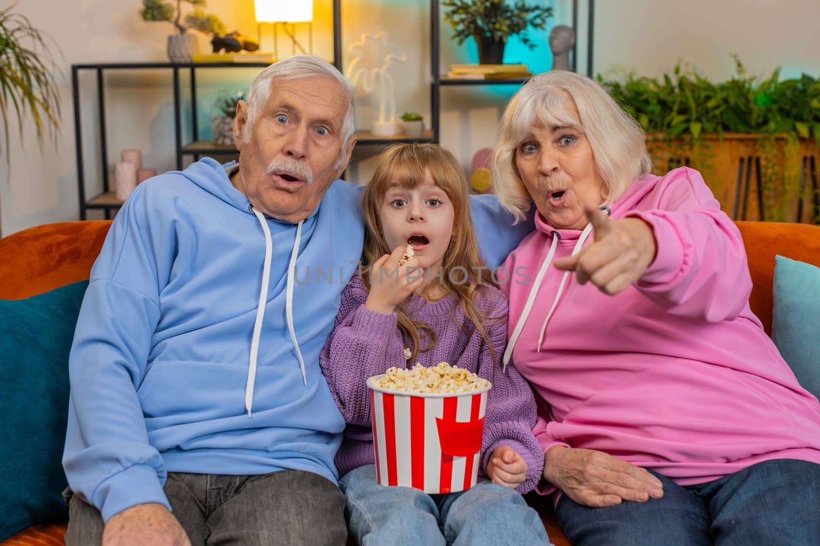 Amazed Caucasian grandfather grandmother and granddaughter eating popcorn and watching movie on sofa at home. Smiling small girl with grandparents enjoying film during weekend in living room apartment