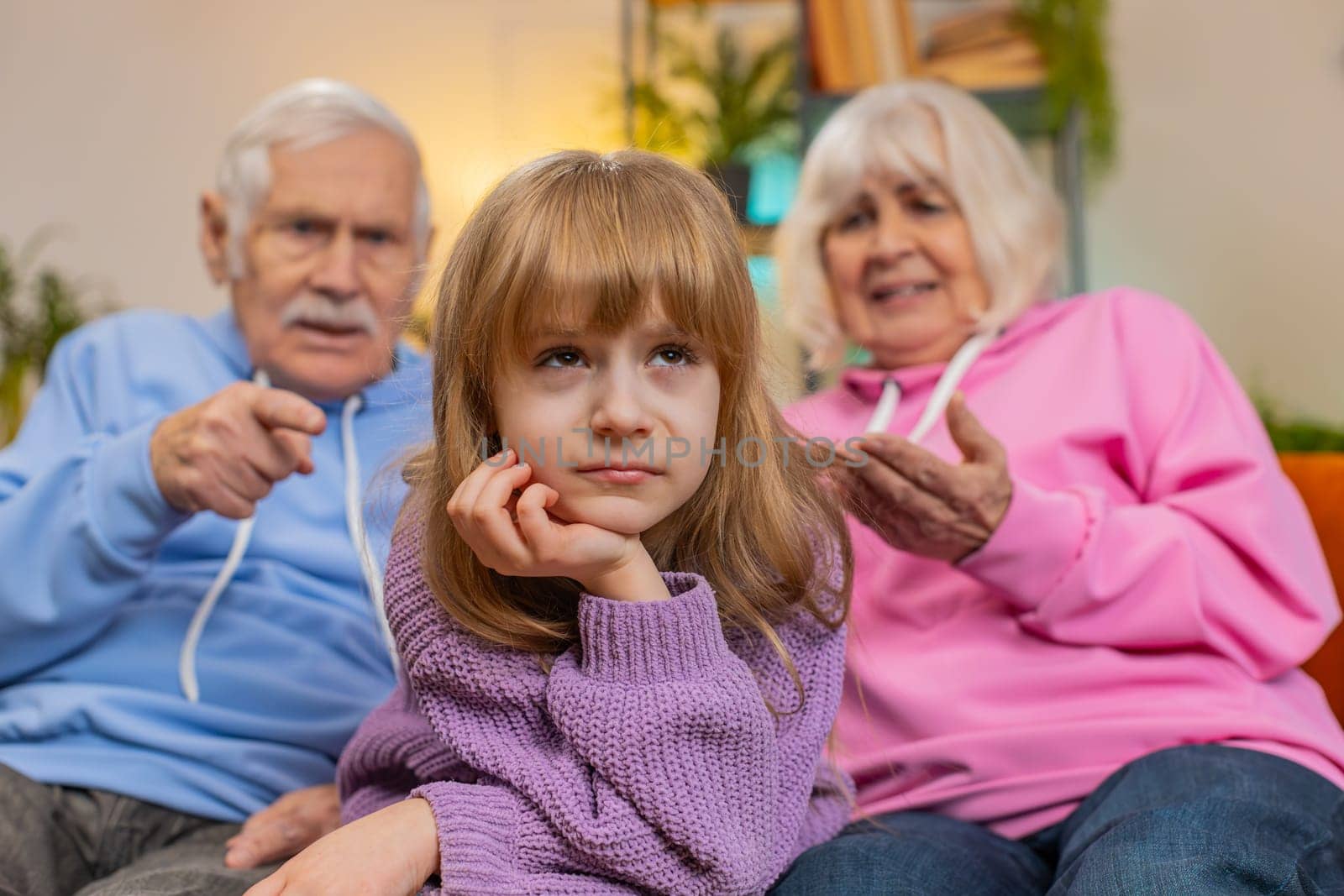 Irritated grandfather and grandmother scolding granddaughter child kid for bad behavior on home sofa by efuror