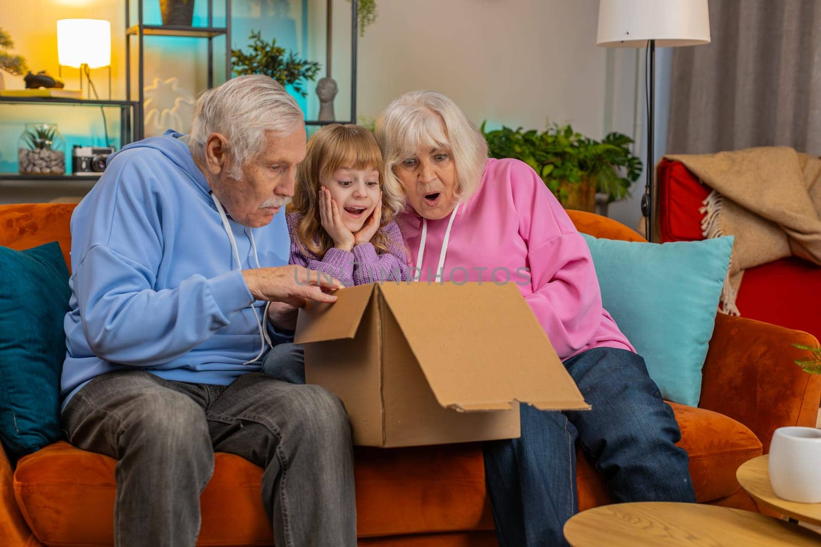 Happy excited Caucasian grandparents and granddaughter girl opening cardboard box together sits on sofa at home. Amazed family consumers unpack good parcel looking inside great purchase delivered.