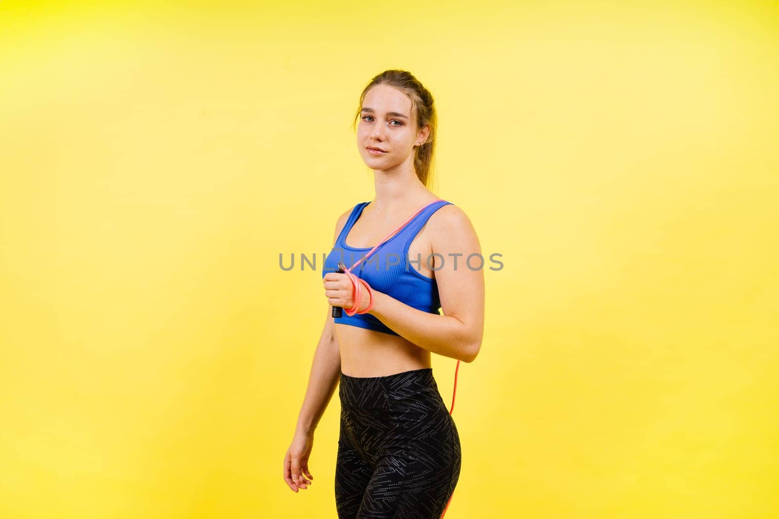 Portrait of gentle muscular woman holding skipping rope on her neck over yellow background