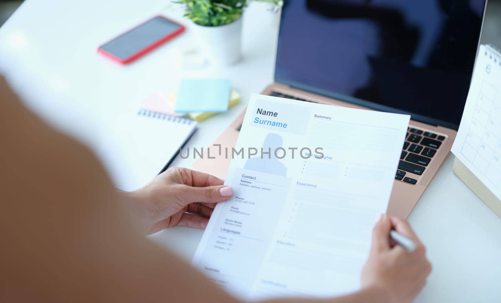Woman sitting at table in front of laptop and holding paper with resume for employment in her hands. Online interview when applying for job concept