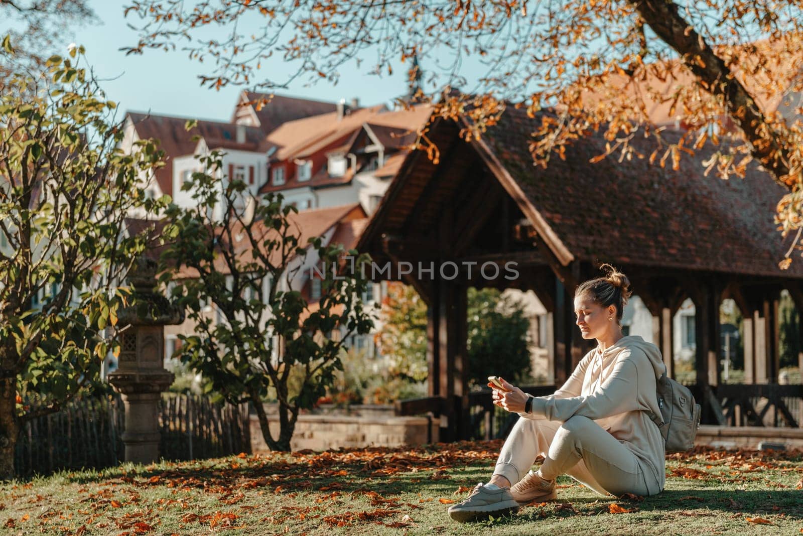 Young fashionable teenage girl with smartphone in park in autumn sitting at smiling. Trendy young woman in fall in park texting. Retouched, vibrant colors. Beautiful blonde teenage girl wearing casual modern autumn outfit sitting in park in autumn. Retouched, vibrant colors, brownish tones.