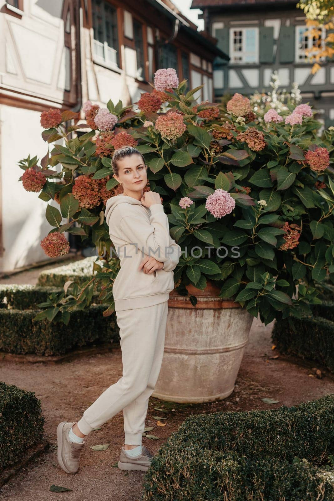 Attractive curly blonde woman walk on the city park street. Girl wear purple hoodie look happy and smiles. Woman make here me gesture standing near pink blooming bush flowers. Happy laughing girl. by Andrii_Ko