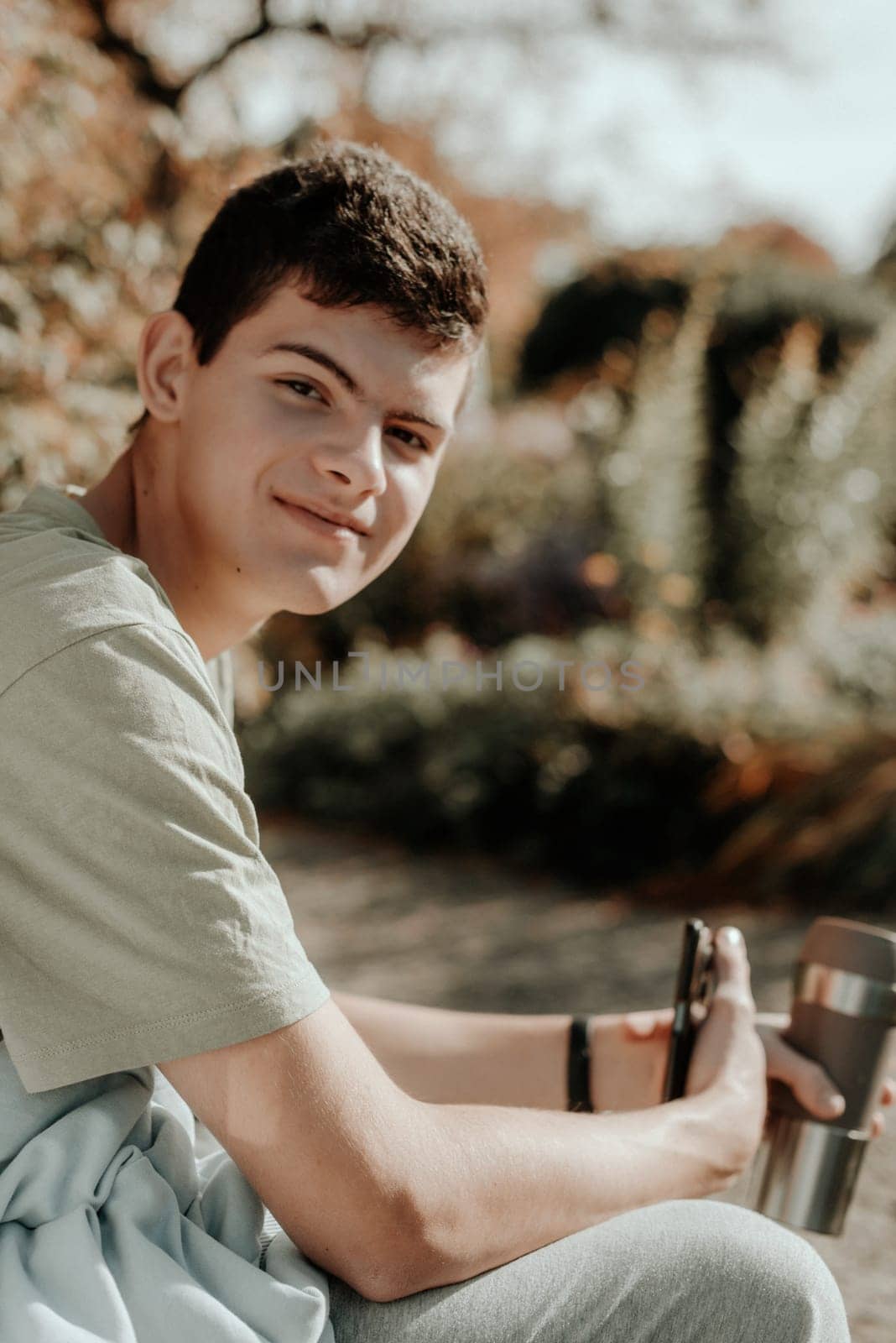 A Teenager Sits On A Bench In The Autumn Park Drinks Coffee From A Thermo Mug And Looks Into A Phone. Portrait Of Handsome Cheerful Guy Sitting On Bench Fresh Air Using Device Browsing Media Smm Drinking Latte Urban Outside Outdoor by Andrii_Ko