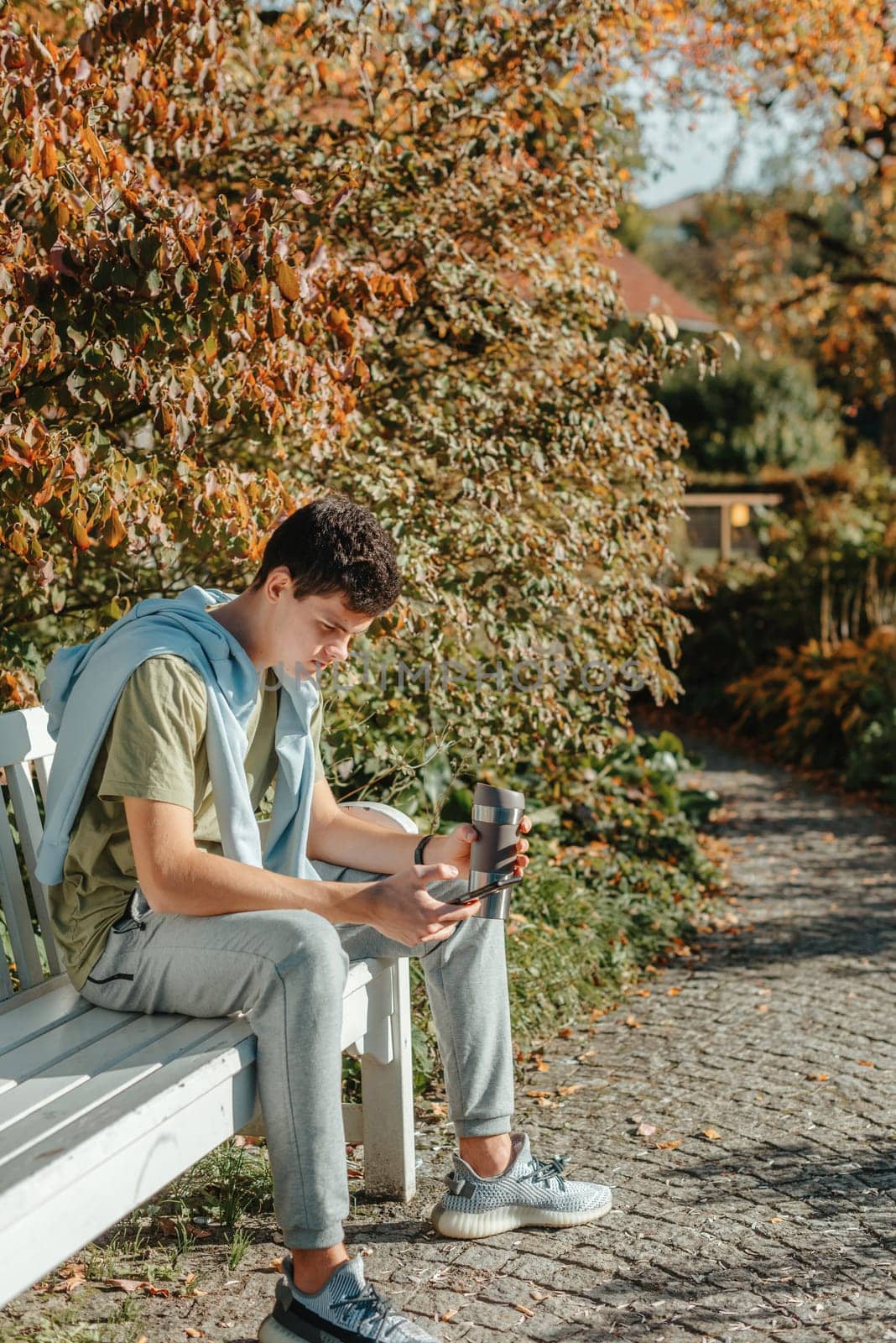 A Teenager Sits On A Bench In The Autumn Park Drinks Coffee From A Thermo Mug And Looks Into A Phone. Portrait Of Handsome Cheerful Guy Sitting On Bench Fresh Air Using Device Browsing Media Smm Drinking Latte Urban Outside Outdoor by Andrii_Ko