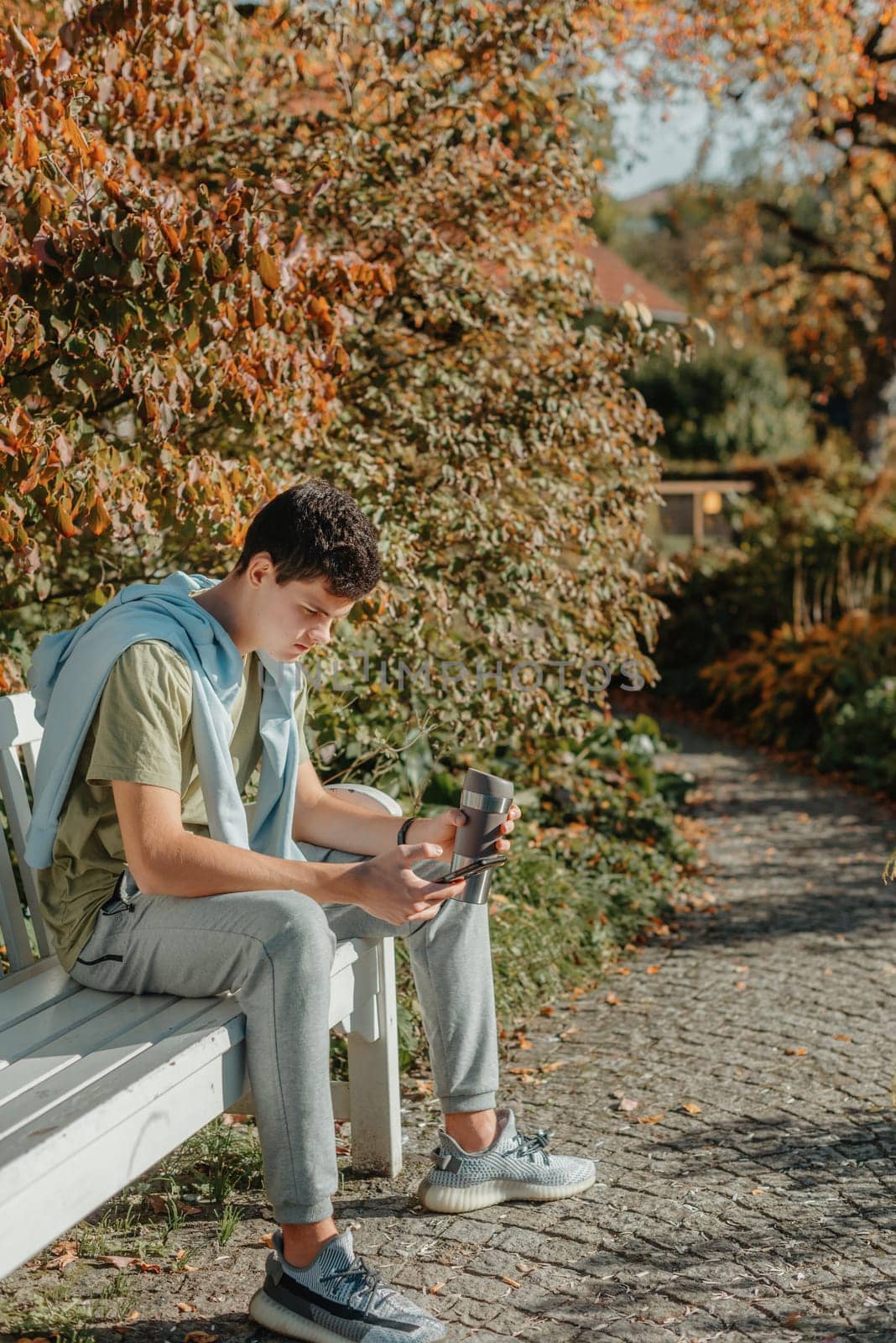 A Teenager Sits On A Bench In The Autumn Park Drinks Coffee From A Thermo Mug And Looks Into A Phone. Portrait Of Handsome Cheerful Guy Sitting On Bench Fresh Air Using Device Browsing Media Smm Drinking Latte Urban Outside Outdoor by Andrii_Ko