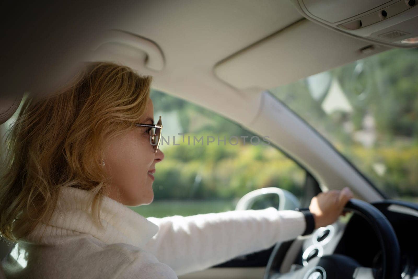 A blonde woman in a white sweater and jeans is driving. Happy woman sitting in a car with a white interior. by Matiunina