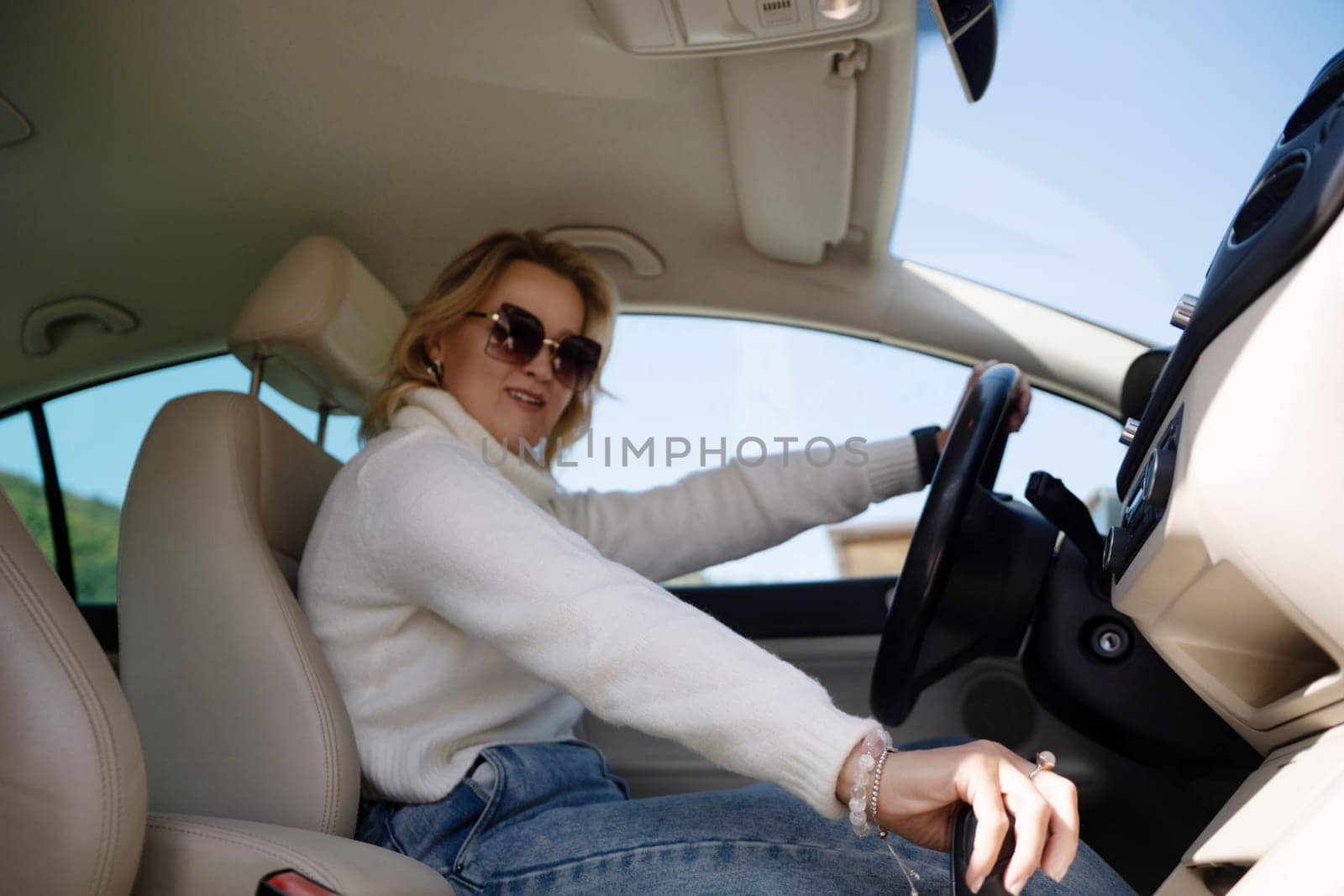 A blonde woman in a white sweater and jeans is driving. Happy woman sitting in a car with a white interior