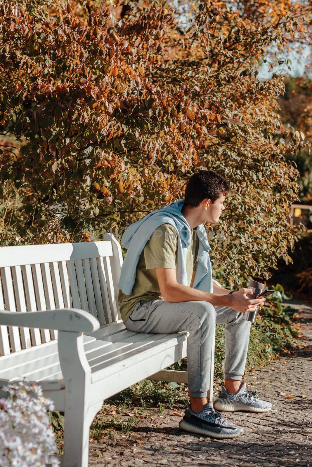 A Teenager Sits On A Bench In The Autumn Park Drinks Coffee From A Thermo Mug And Looks Into A Phone. Portrait Of Handsome Cheerful Guy Sitting On Bench Fresh Air Using Device Browsing Media Smm Drinking Latte Urban Outside Outdoor by Andrii_Ko