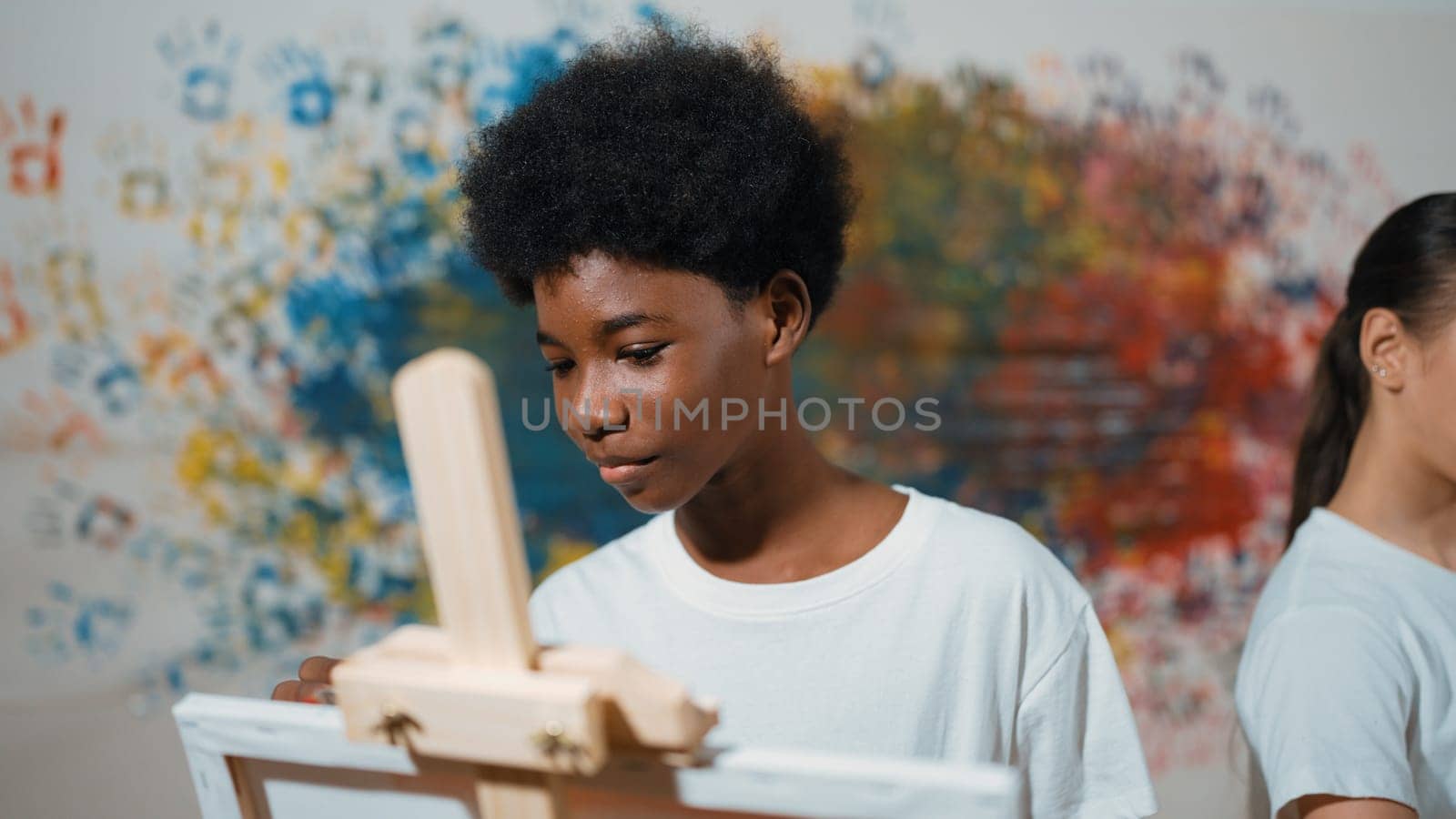 African boy painting canvas with watercolor while happy caucasian girl draw cool tone picture at colorful stained wall. Multicultural highschool student attend creative activity together. Edification.