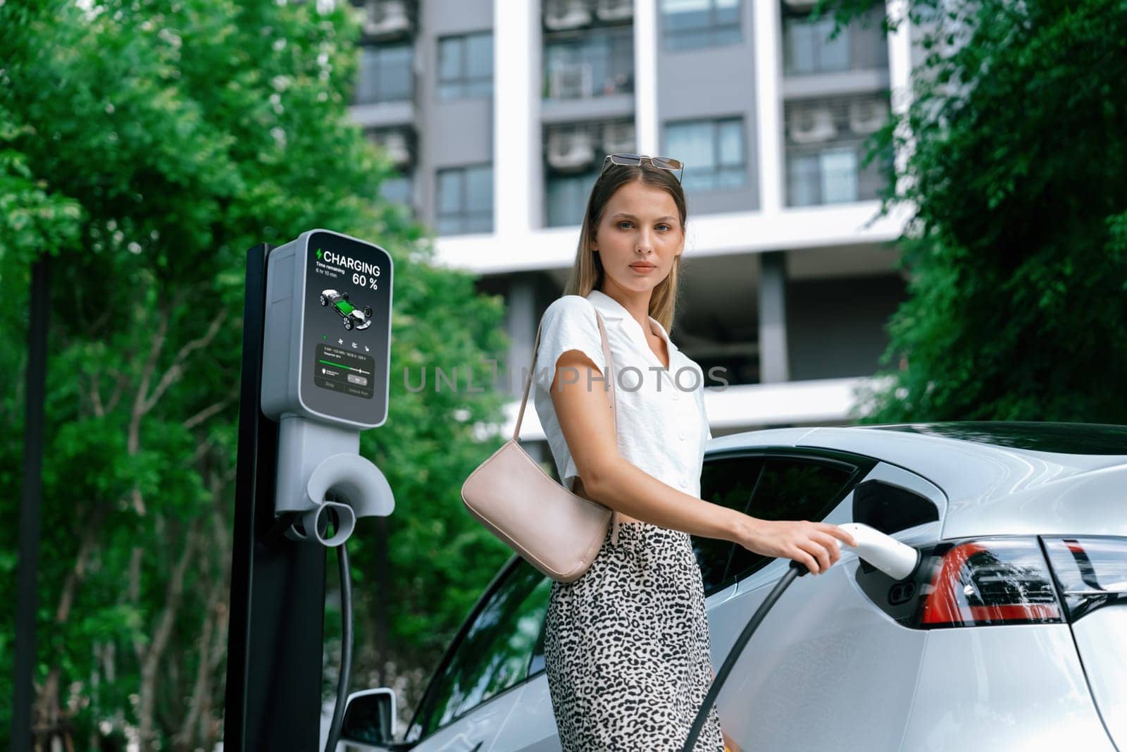 Young woman travel with EV electric car charging in green sustainable city outdoor garden in summer. Urban sustainability lifestyle by green clean rechargeable energy of electric BEV vehicle innards