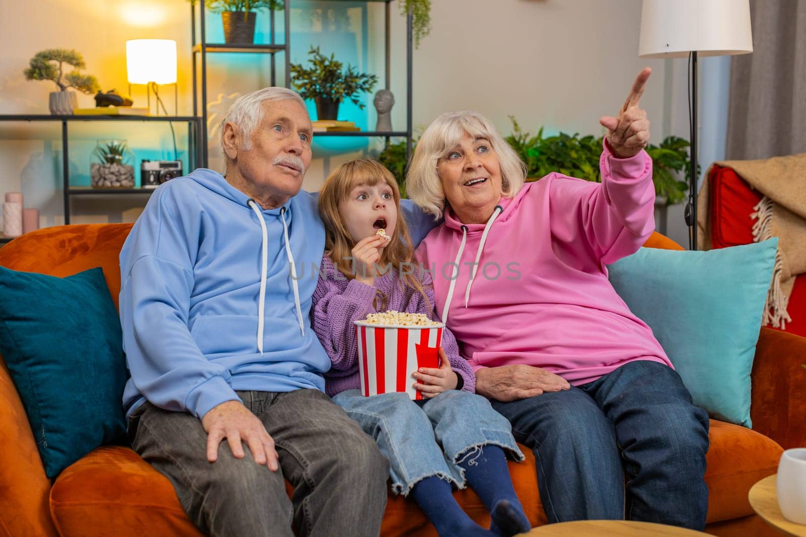 Happy Caucasian grandfather, grandmother and granddaughter eating popcorn and watching movie on sofa at home. Smiling small girl with grandparents enjoying film during weekend in living room apartment