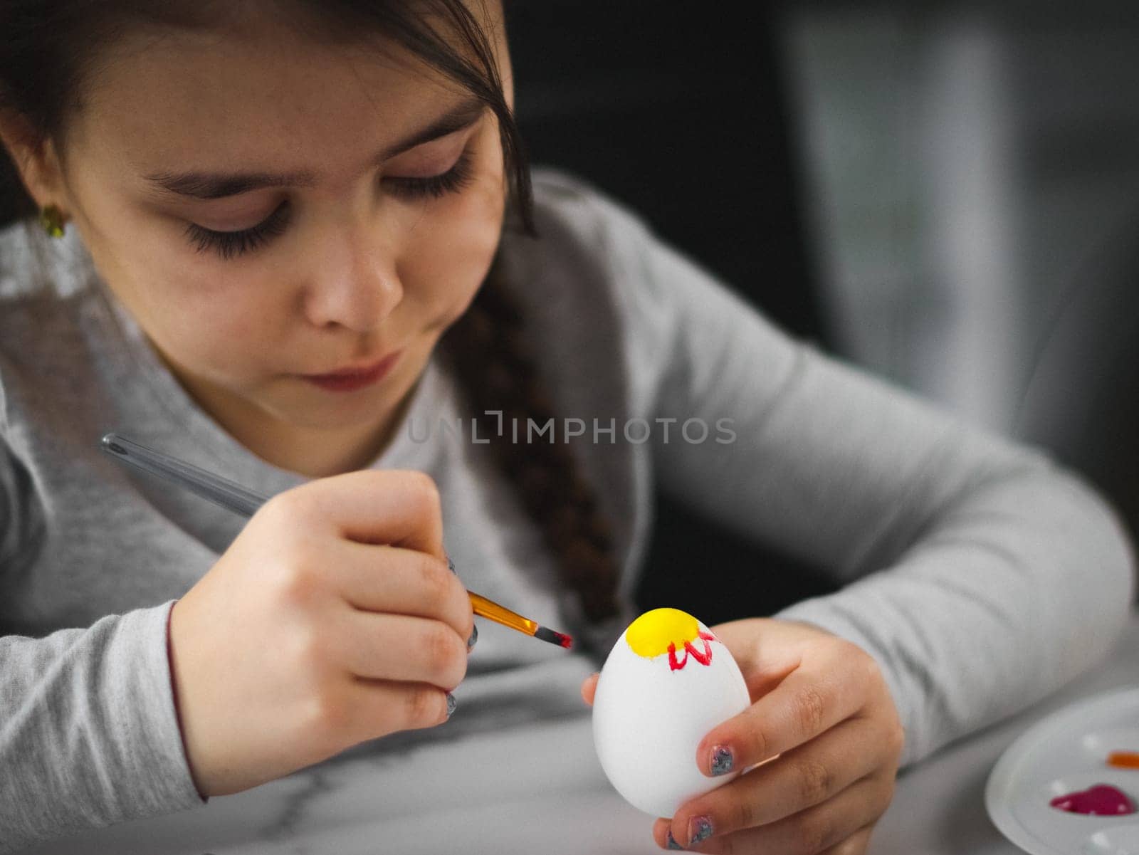 A little caucasian girl in a gray turtleneck enthusiastically paints an Easter egg with a brush with acrylic paint at a marble table with a palette, willow branches, eggs in a box for diy preparation for the Easter holiday, close-up side view. The concept of crafts, needlework, artisanal, children art, diy, at home.