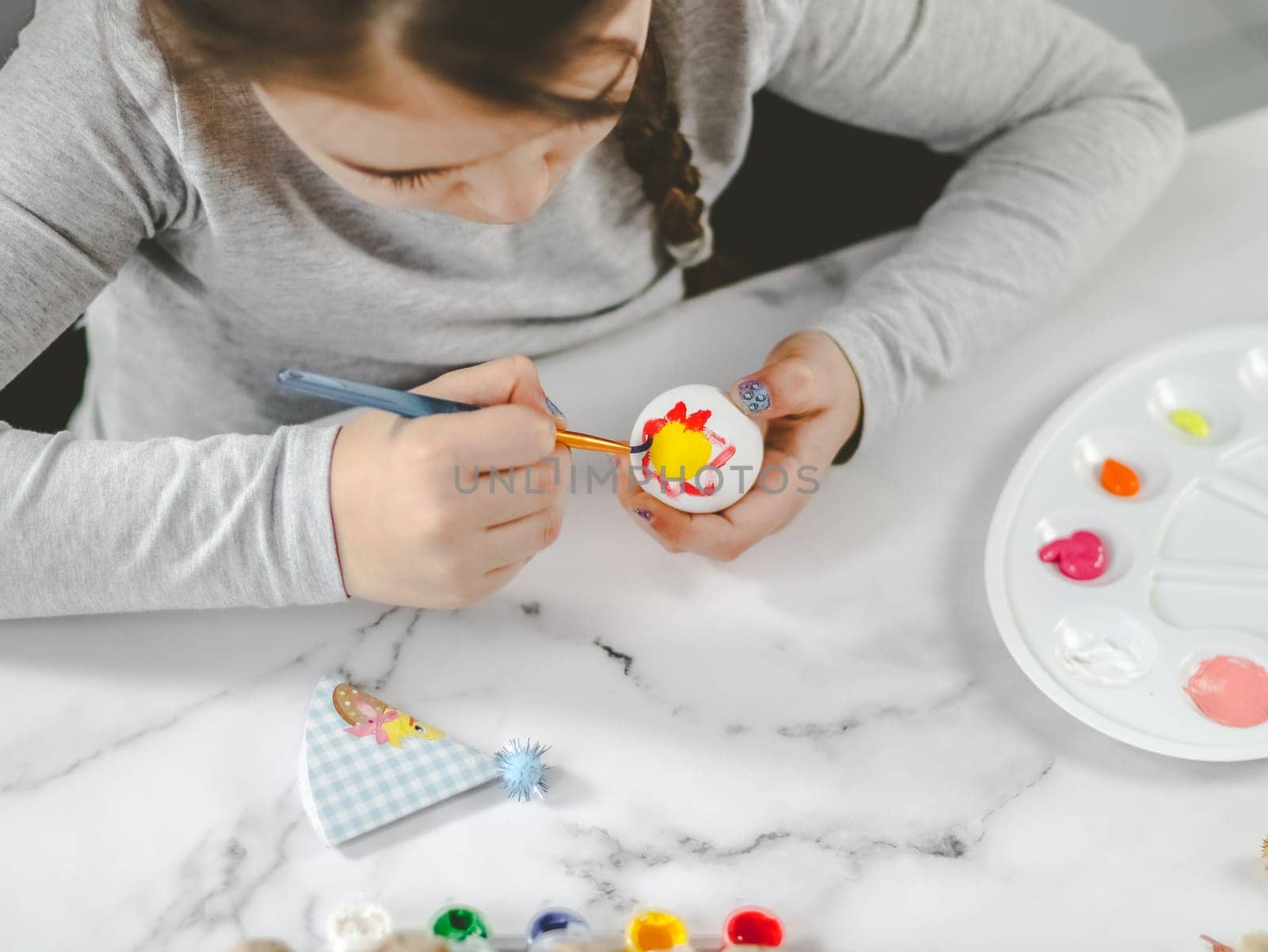 Little caucasian girl in a gray turtleneck enthusiastically paints an Easter egg with a brush of red-yellow acrylic paint at a marble table with a palette, willow branches, eggs in a box for diy preparation for the Easter holiday, close-up top view. The concept of crafts, needlework, at home,diy,artisanal,children art,children creative.