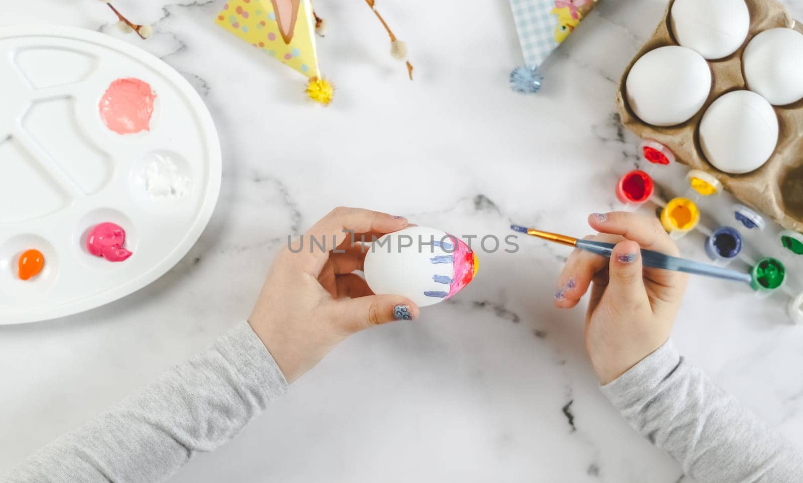 Little caucasian girl paints an easter egg with a brush with blue stripes of acrylic paint, sitting at a marble table with a palette for diy preparation for the easter holiday, flat lay close-up. The concept of crafts, needlework, at home, children art, children creative.
