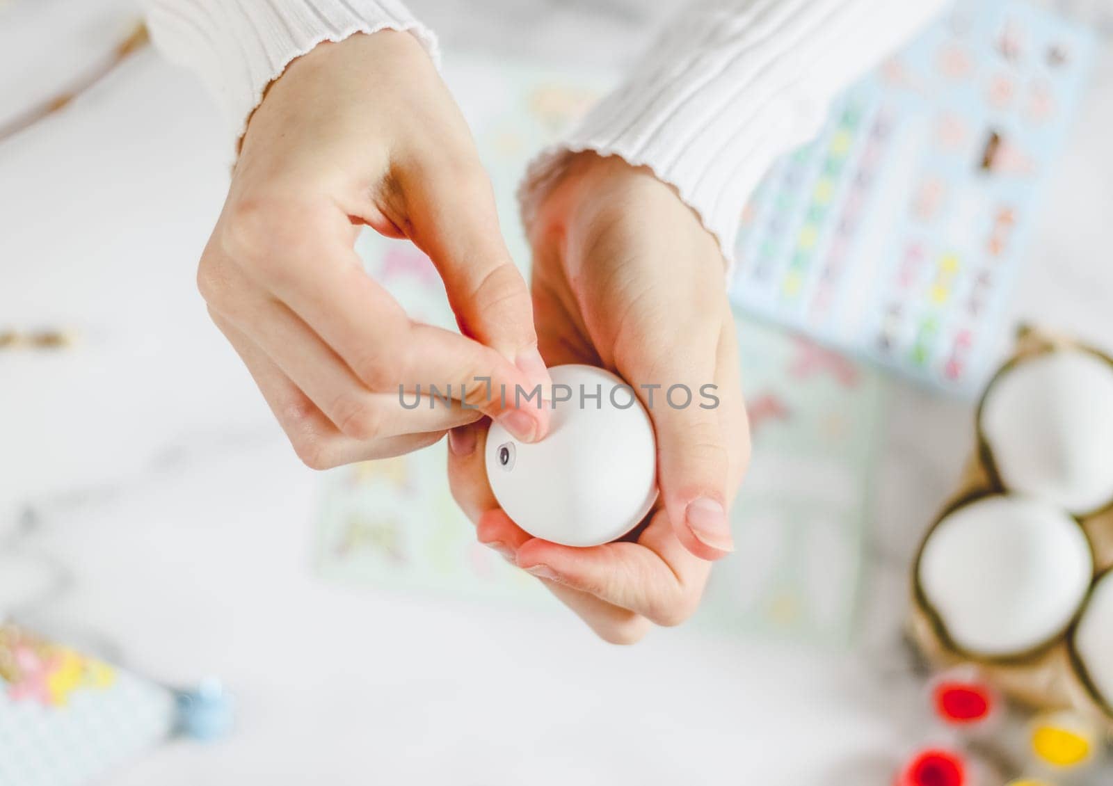 Children's hands of a caucasian teenager girl in a white turtleneck hold one white egg and stick adhesive decorative eyes, sit at a marble table with tassels, prepare crafts for the Easter holiday, flat lay close-up. Craft concept, handicraft, at home, children art, diy.