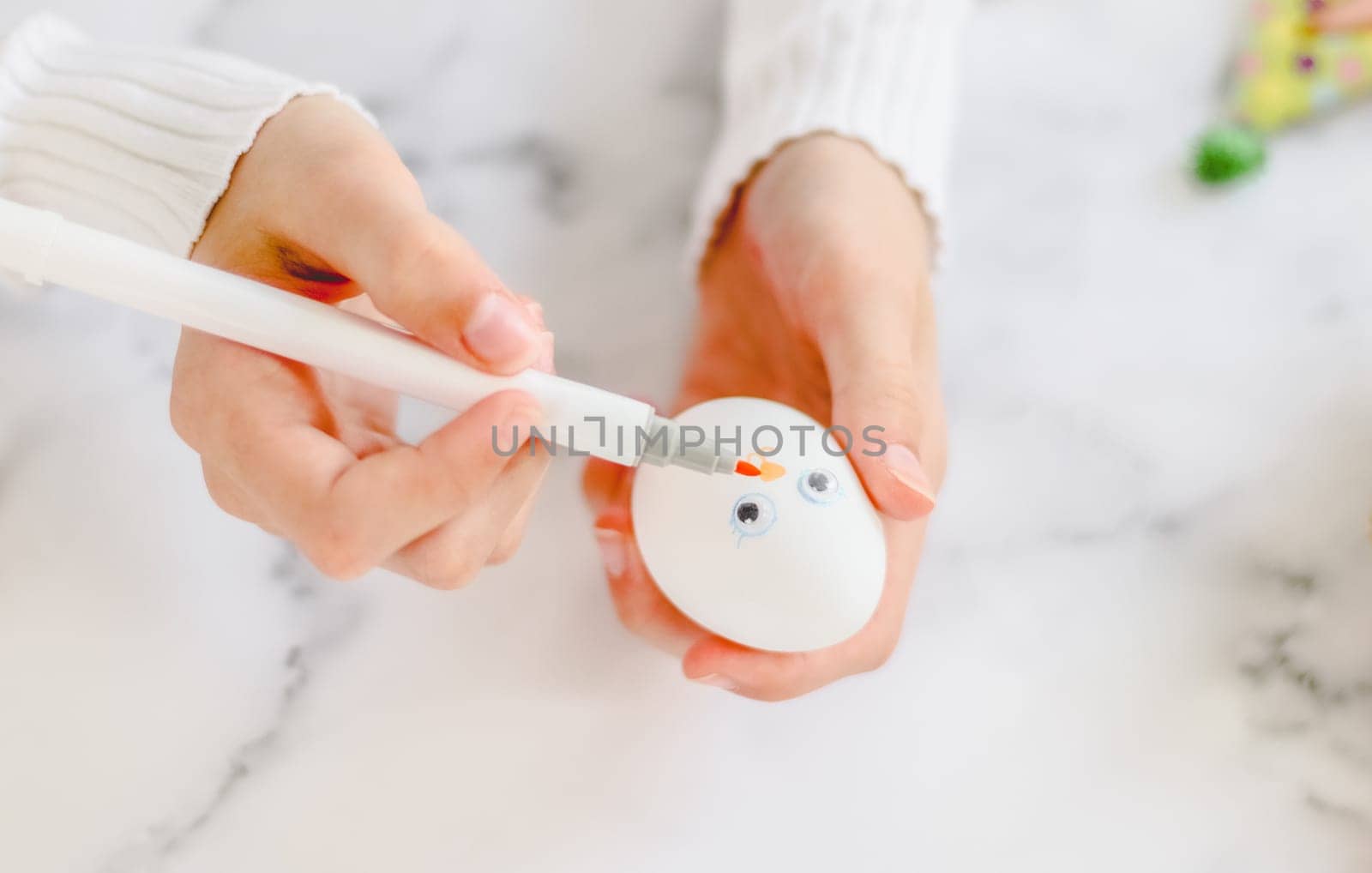 The hands of a caucasian girl in a white turtleneck draw with a marker on an egg the muzzle of an easter bunny on a marble table for diy preparation for the easter holiday, close-up top view. The concept of crafts, needlework, minimalism,artisanal,at home,children art,children creative.