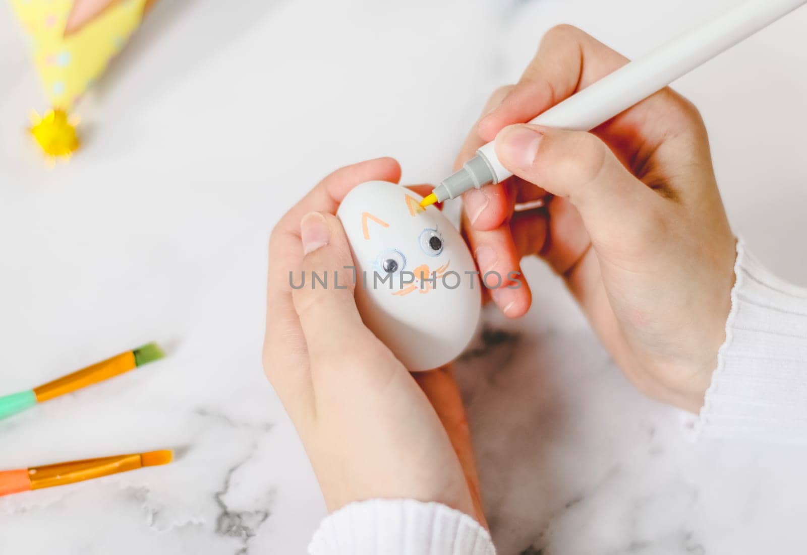 The hands of a caucasian girl in a white turtleneck draw with an orange marker on an egg the ears of an easter bunny on a marble table to prepare for the easter holiday, close-up flat lay. The concept of craft, needlework, at home, artisanal, children creative, easter preparation, children creative.