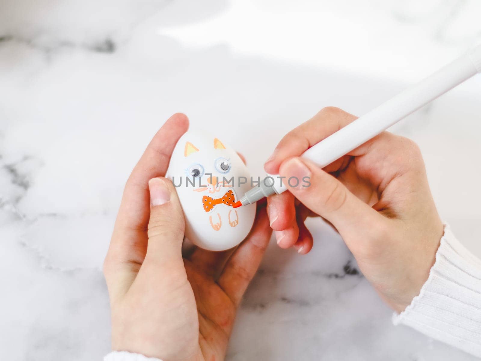 Hands of a caucasian teenage girl in a white turtleneck holding an easter egg and a marker drawing bunny paws while sitting at a marble table, top view close-up. The concept of crafts, needlework, at home, diy, children art, diy, Easter preparation, children creative.