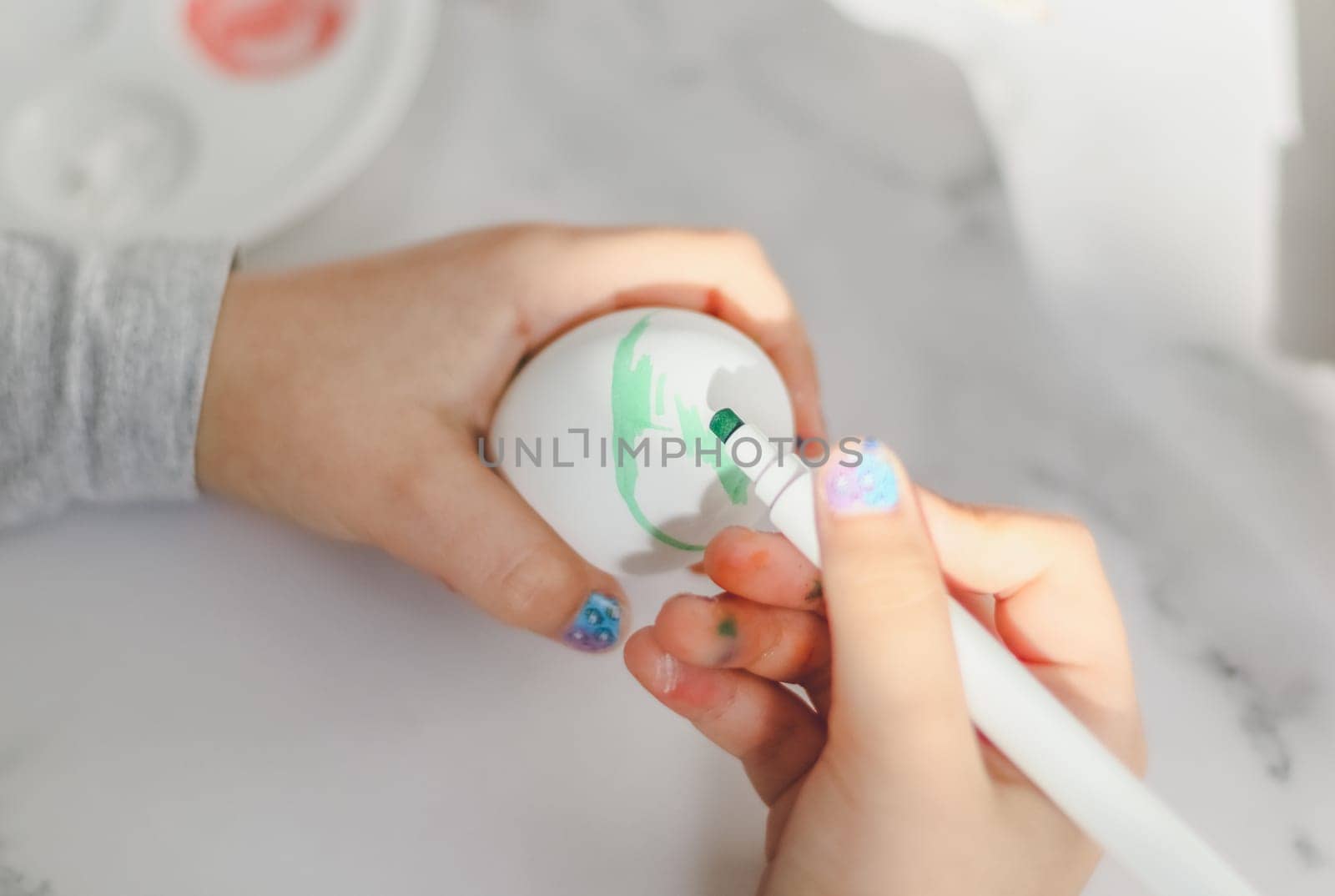 The hands of a caucasian girl in a gray turtleneck draw with a green marker on a white egg, sit at a marble table with a palette and acrylic paints, close-up side view. The concept of crafts, needlework, children's creativity, preparation for easter, children's creativity, home, crafts.