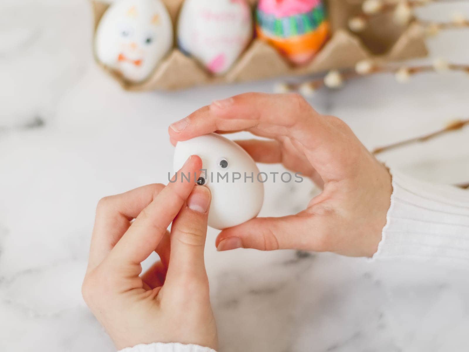 Children's hands hold one white egg and glue eye stickers, sit at a marble table with depth of field, prepare crafts for the Easter holiday, close-up side view. The concept of craft, needlework, at home, children creative, diy, crafts, children art.