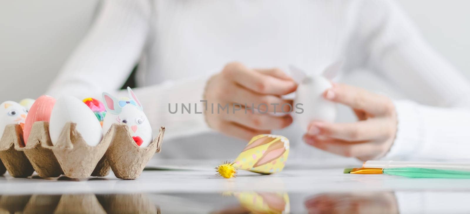 Cardboard box with easter bunny egg craft and caucasian teenage girl in white turtleneck behind needlework process in blur sitting at marble table with reflection from below, close up side view. The concept of craft, needlework, at home, diy, children art, artisanal, children's creative, step by step, diy.