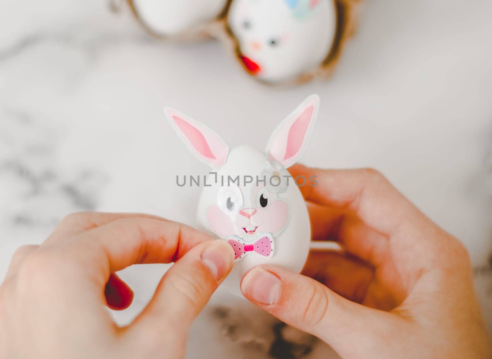 Hands of a caucasian girl pasting bow stickers on an easter bunny egg with ears, sitting at a marble table with depth of field, preparing crafts for the easter holiday, close-up side view. The concept of craft, needlework, at home, diy,children art,artisanal,children creative,step by step.