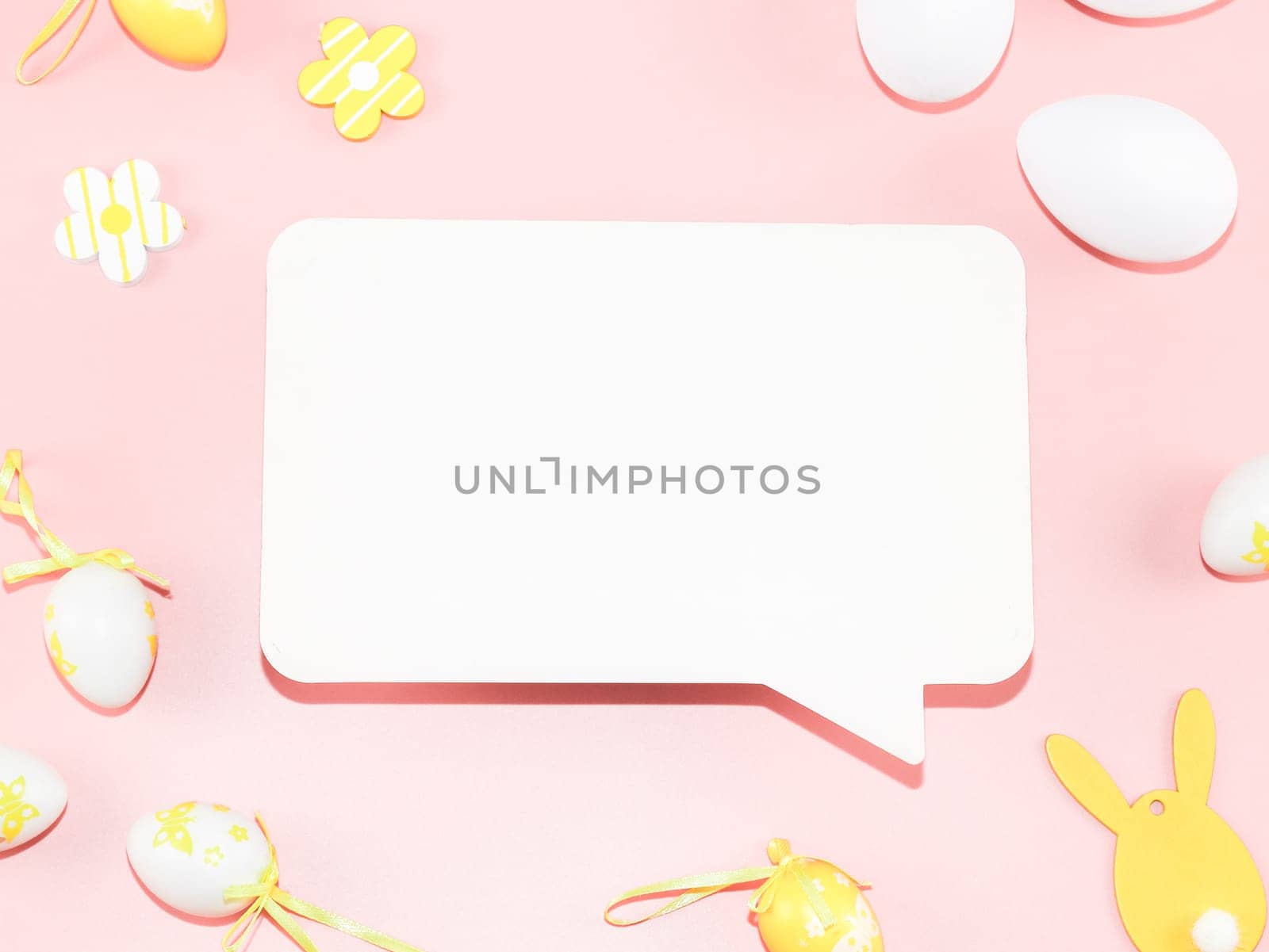 Empty white callout, decorative yellow-white eggs, and a wooden easter bunny lie on a light pink background with copy space for your tex, flat lay close-up. Happy easter concept, holiday banner, blank.