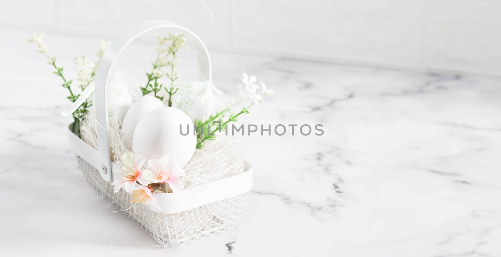 Decorative white eggs in a metal basket with eggs, spring flowers and straw stand on the left on a marble table with copy space on the right in the early morning, close-up side view. Happy easter concept, holiday banner.