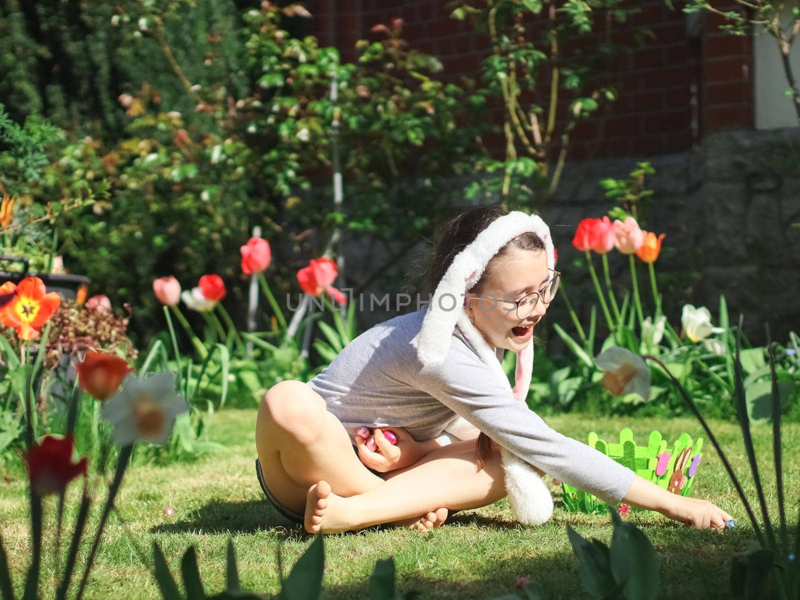 Caucasian beautiful happy girl with bunny ears on her head smiles chocolate easter eggs while sitting on the lawn in the backyard of the house, close-up side view. Easter tradition concept, Easter celebration.