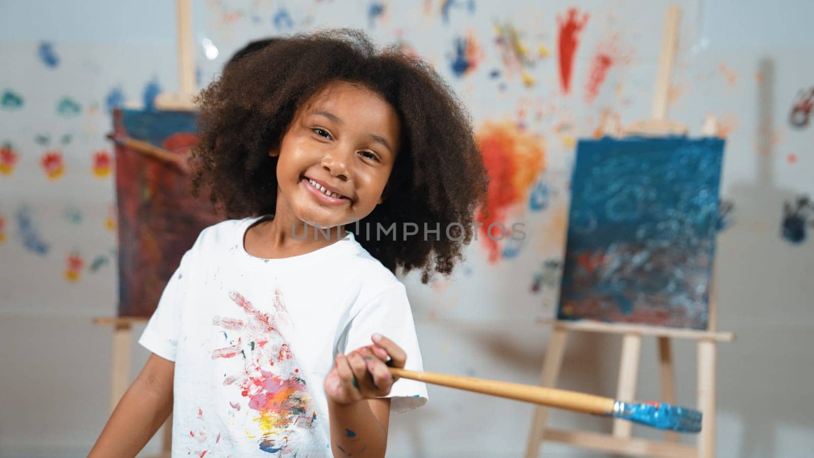 Cute african girl holding painted brush while student drawing canvas behind. Multicultural children attend in art lesson while pretty learner looking at camera. Creative activity concept. Erudition.