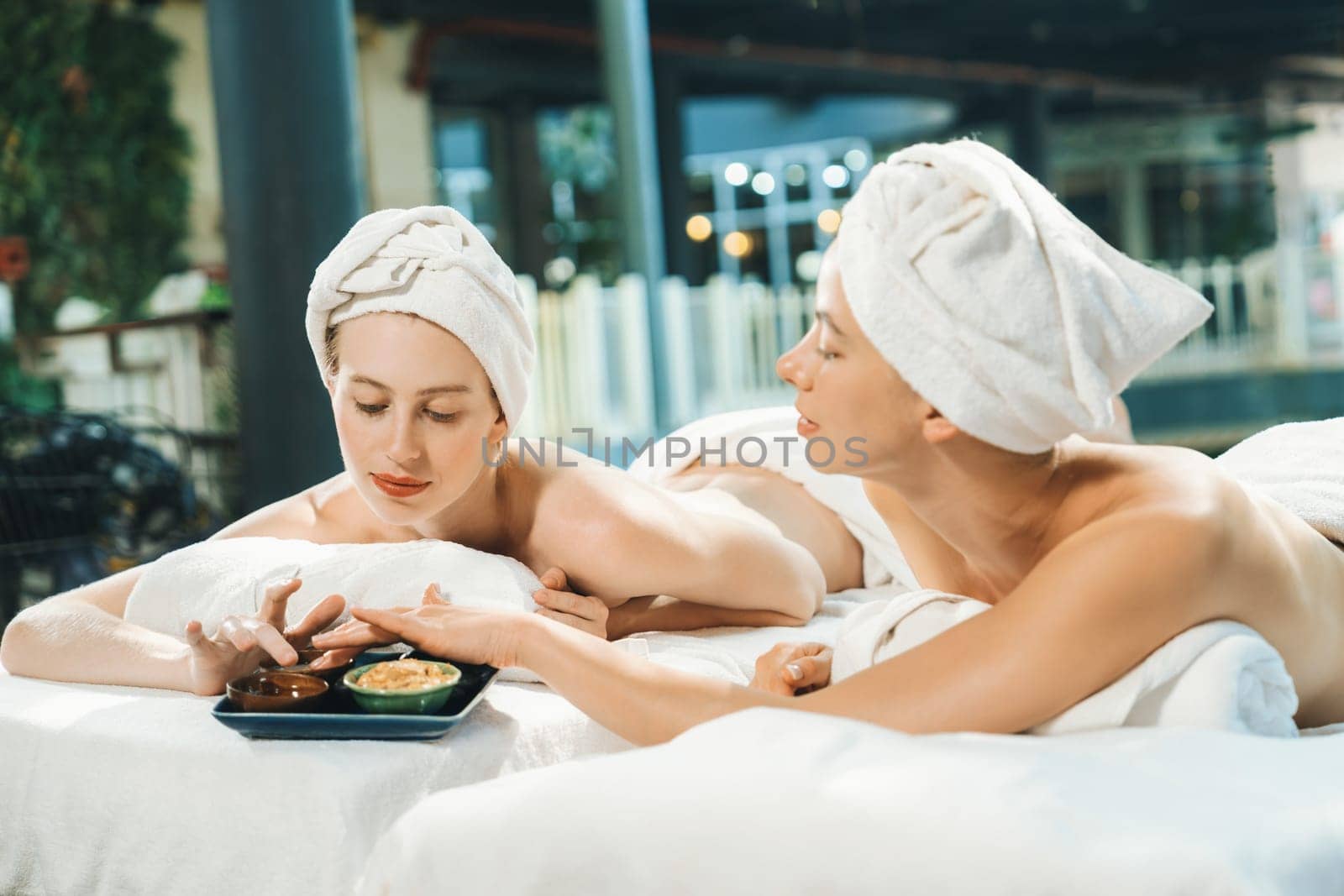 Couple of beautiful young girls lie on spa bed during interested in homemade beauty facial mask. Attractive woman touching herbal facial mask. Surrounded with nature environment. Tranquility.