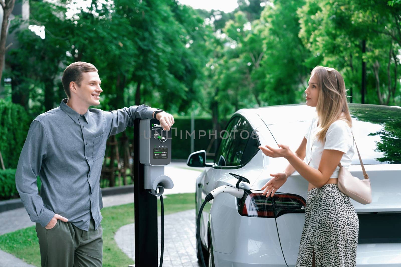 Young couple travel with EV electric car charging in green sustainable city outdoor garden in summer shows urban sustainability lifestyle by green clean rechargeable energy of electric vehicle innards