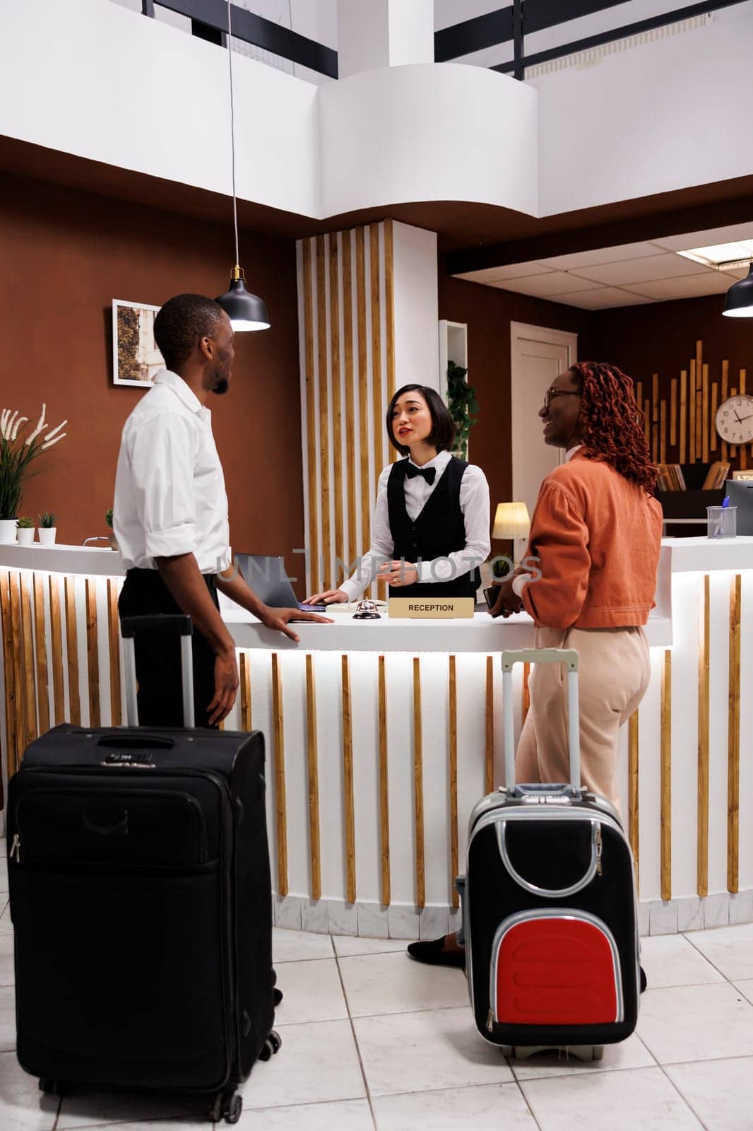 Asian woman welcoming couple at hotel, reviewing guests information before doing check in process to show room. Receptionist registering people arriving at resort, front desk.