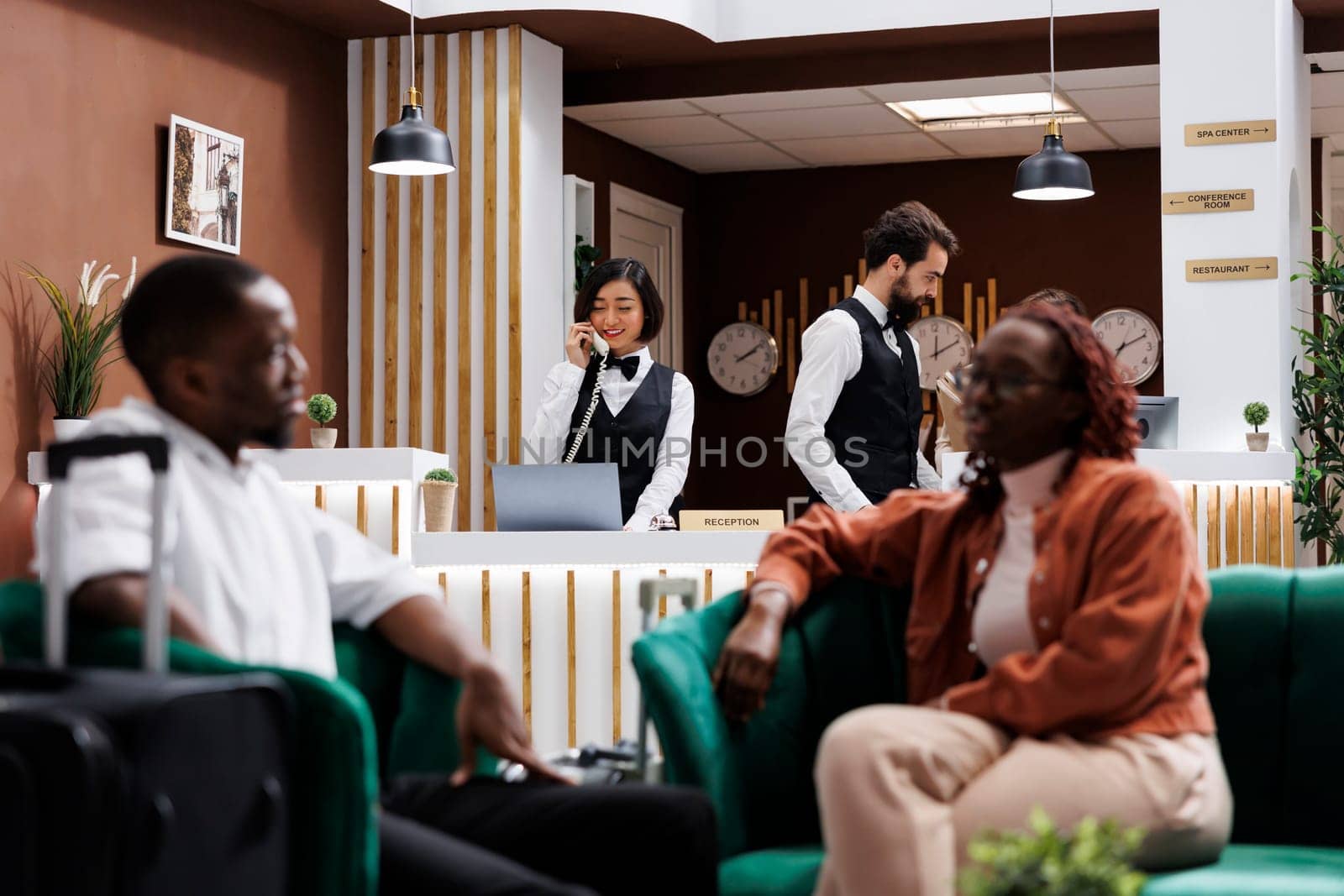 Employees working at front desk in hotel lobby, checking records and room availability at reception. Two receptionists answering landline phone call to make online reservations.