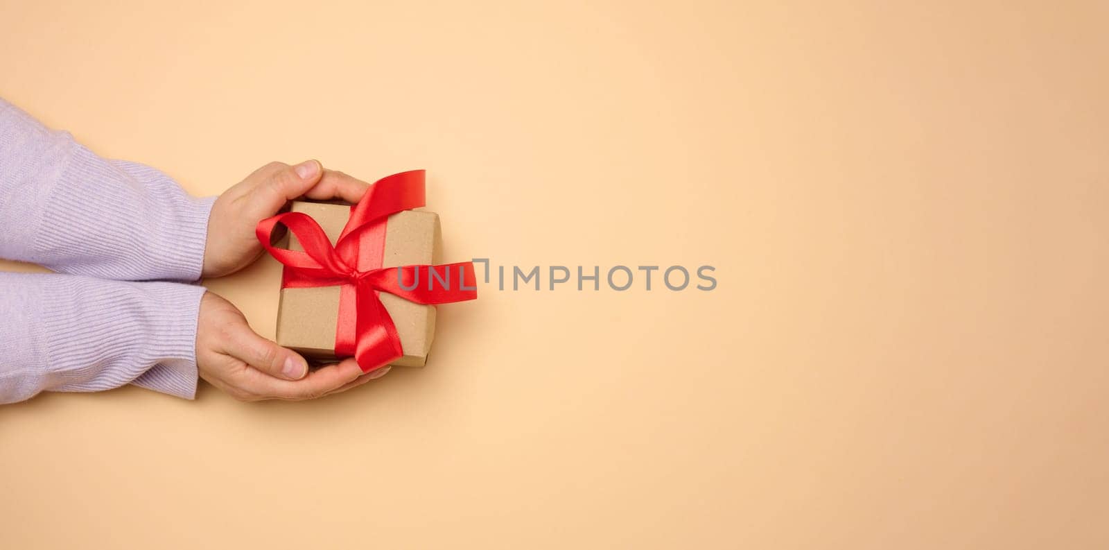 A woman's hand holds a gift box wrapped in a red silk ribbon on a beige background, top view by ndanko