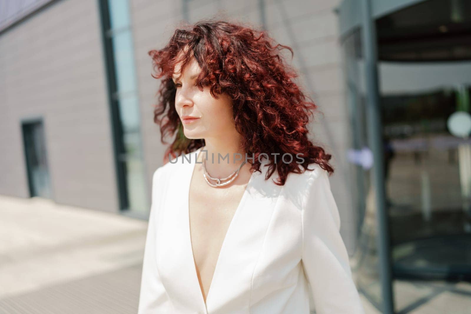 Portrait of a woman standing near a supermarket building. Caucasian model with long dark hair, wearing a white jacket and colored trousers